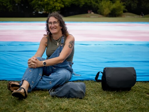 A woman with dark curly hair, in a denim jumpsuit, sitting on grass in front of a giant trans flag, layed out on the grass behind her